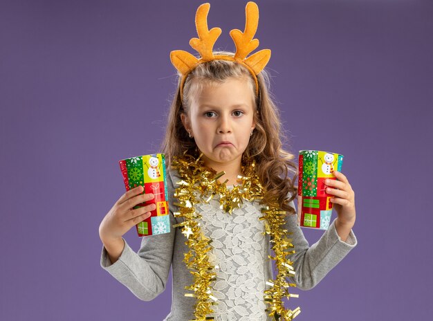 Sad little girl wearing christmas hair hoop with garland on neck holding christmas cups isolated on blue background