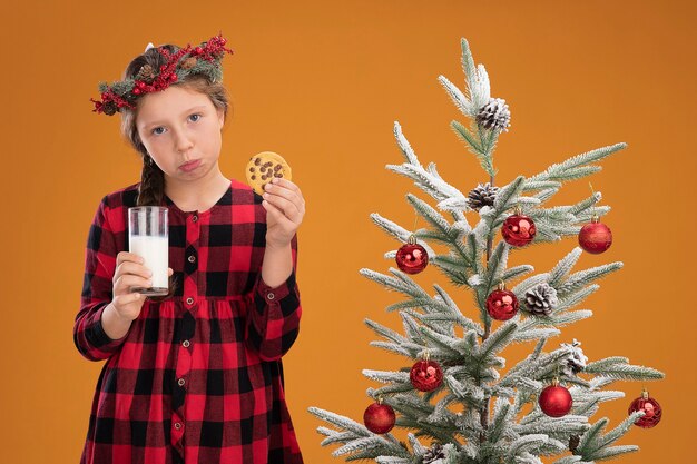 顔に悲しい表情で探している牛乳とクッキーのガラスを保持しているチェックシャツにクリスマスリースを着ている悲しい少女