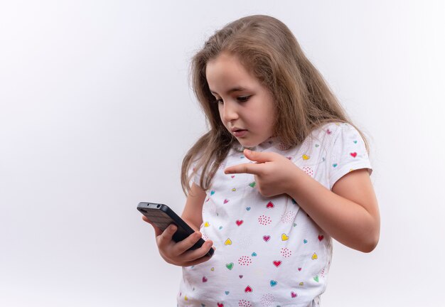 Грустная маленькая школьница в белой футболке указывает на телефон на изолированном белом фоне