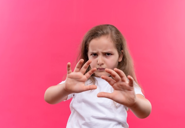Грустная маленькая школьница в белой футболке показывает жест стоп на изолированном розовом фоне