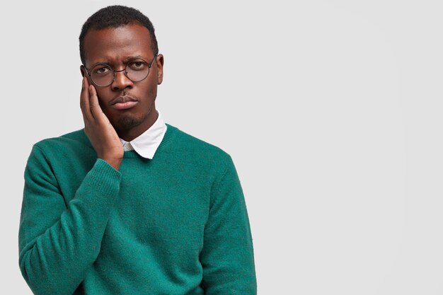 Sad lonely dissatisfied young black man keeps hand on cheek, feels displeased
