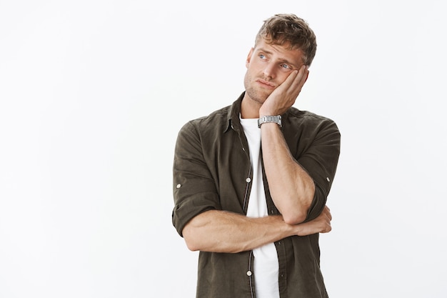 Sad and lonely moody cute european masculine man with beard and blond hair leaning head on palm from boredom looking outside turning left and gazing indifferent with sorrow over gray wall