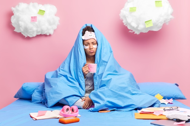 Sad lonely woman wrapped in soft blanket holds mug of coffee with stickers