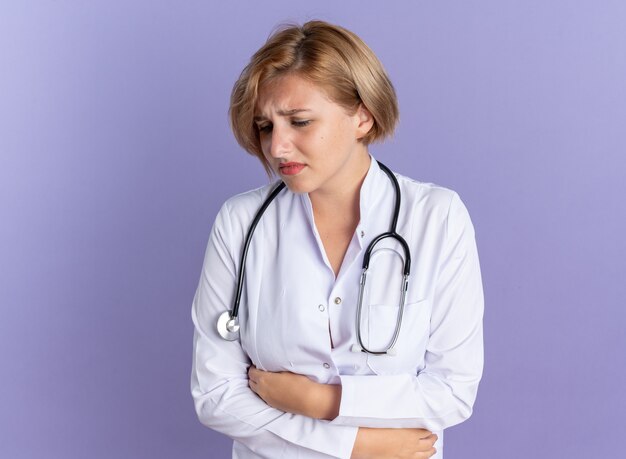 聴診器で医療ローブを身に着けている若い女性医師を見下ろす悲しいは、青い背景で隔離の痛む胃をつかんだ