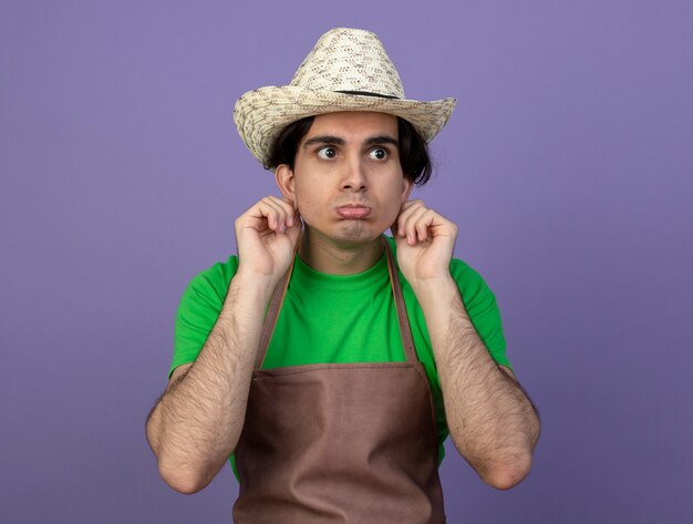 Sad looking at side young male gardener in uniform wearing gardening hat holding ears isolated on purple