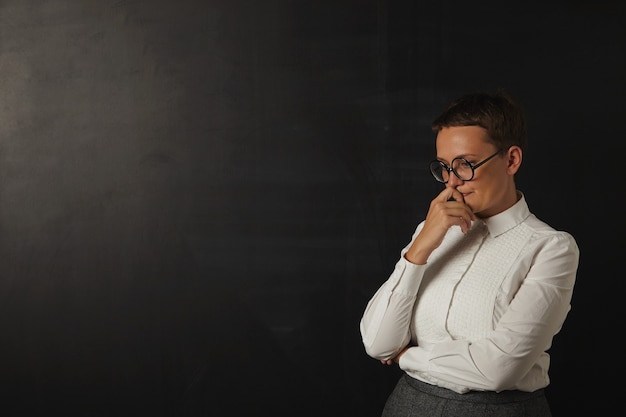 Sad looking young female teacher in white blouse and gray skirt deep in thought next to a blank blackboard