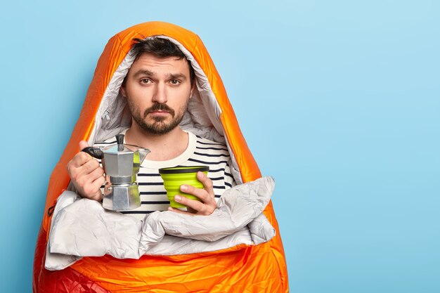 Sad male camper prepares fresh drink, holds coffee pot, spends free time in nature, enjoys summer morning, wrapped in sleeping bag, stands against blue wall