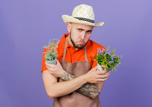 Sad male gardener wearing gardening hat crosses arms holding flowerpots