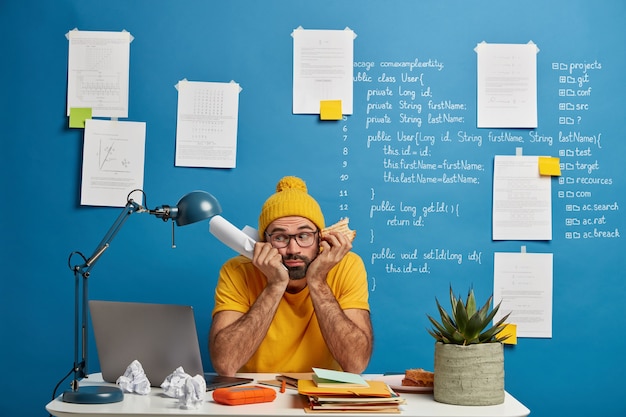 Free photo sad male student preparess to write exam testing, poses at coworking space, holds paper and burger, wears yellow clothes