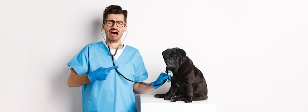 Бесплатное фото Печальный мужчина-ветеринар осматривает симпатичного маленького собачьего мопса со стетоскопом, указывающим на домашнее животное и плачущим стоя