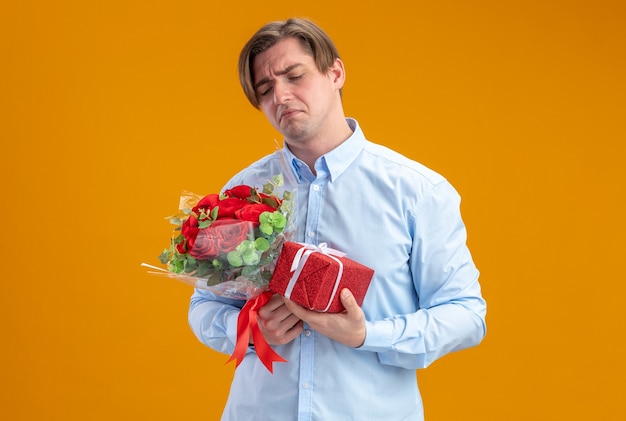 грустный мужчина в синей рубашке держит букет красных роз и презрительно сжимает губы с разочарованным выражением дня святого валентина, стоящим над оранжевой стеной