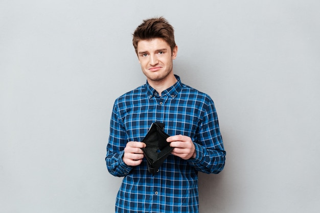 Free photo sad man standing over grey wall holding purse without money.