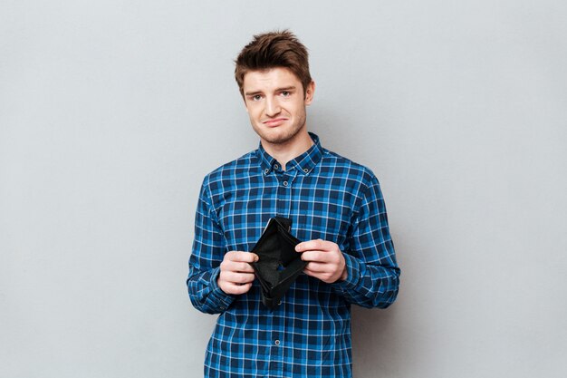 Sad man standing over grey wall holding purse without money.