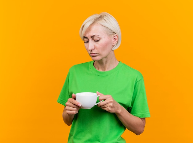 Free photo sad middle-aged blonde slavic woman holding and looking at cup of tea isolated on yellow background with copy space