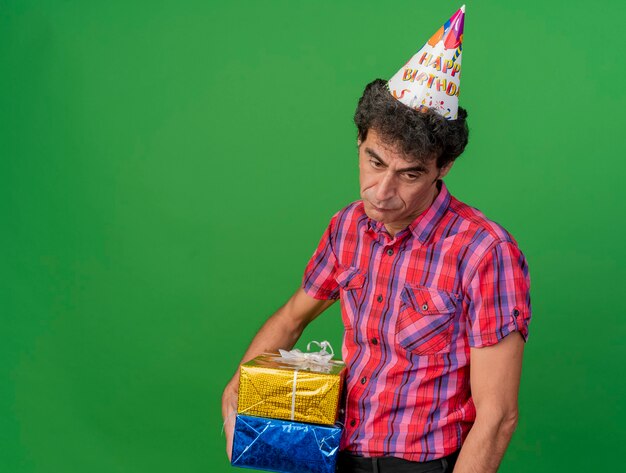 Sad middle-aged caucasian party man wearing birthday cap holding gift packs looking at side isolated on green background with copy space