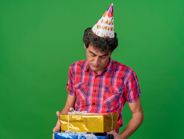 Sad middle-aged caucasian party man wearing birthday cap holding and looking at gift packs isolated on green background with copy space