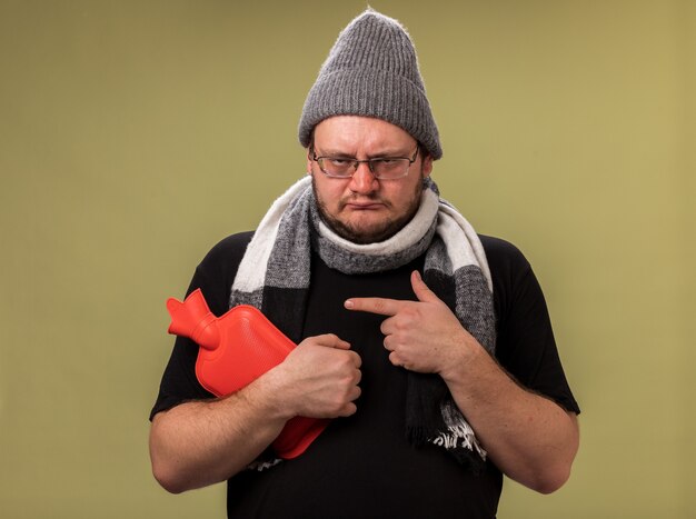 Sad middle-aged ill male wearing winter hat and scarf holding and points at hot water bag isolated on olive green wall