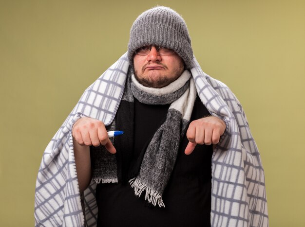 Sad middle-aged ill male wearing winter hat and scarf wrapped in plaid holding thermometer 