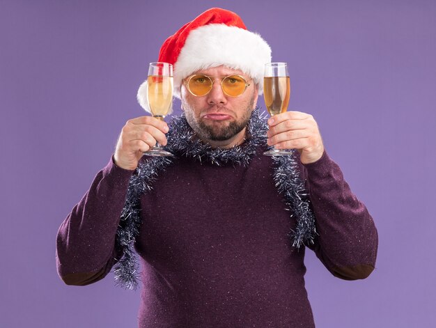 보라색 벽에 고립 된 샴페인 두 잔을 들고 안경 목에 산타 모자와 반짝이 화환을 입고 슬픈 중년 남자
