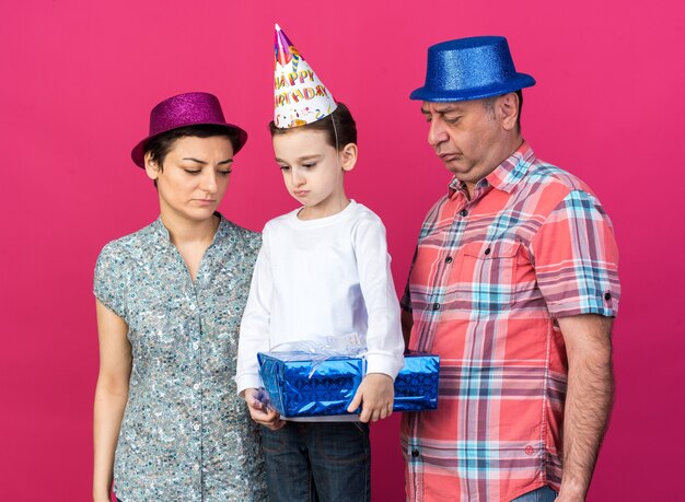 sad mother and father with party hats looking at their disappointed son holding gift box isolated on pink wall with copy space