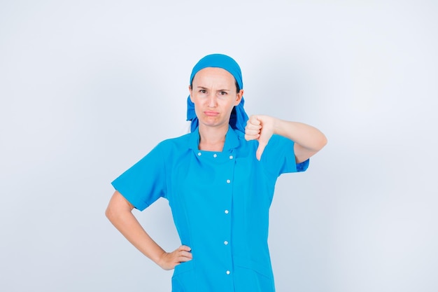 Sad nurse is showing dislike gesture and putting other on waist on white background