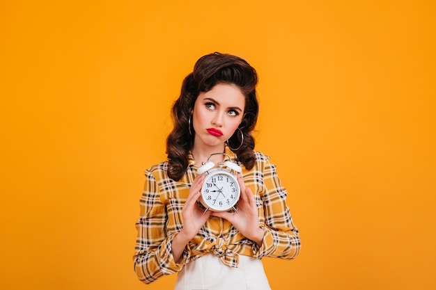 Free photo sad pinup girl holding clock. studio shot of pensive young woman in yellow checkered shirt.