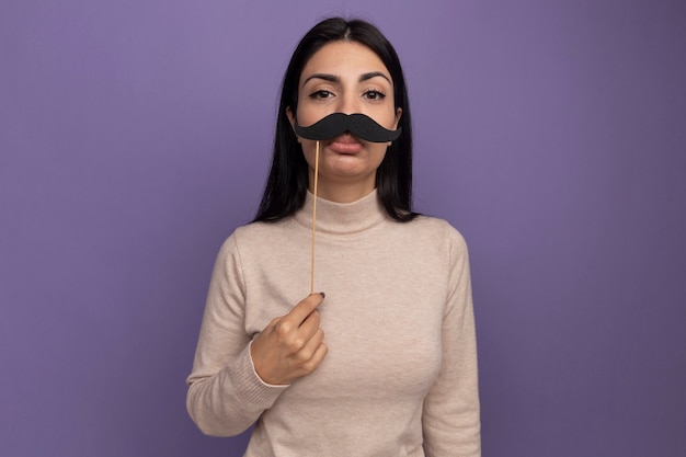 Sad pretty brunette caucasian girl holds fake mustache on stick on purple