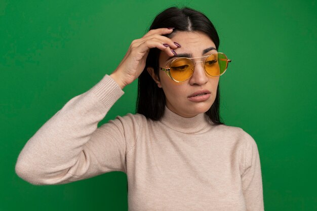 Грустная симпатичная брюнетка кавказская девушка в солнцезащитных очках кладет руку на лоб на зеленый