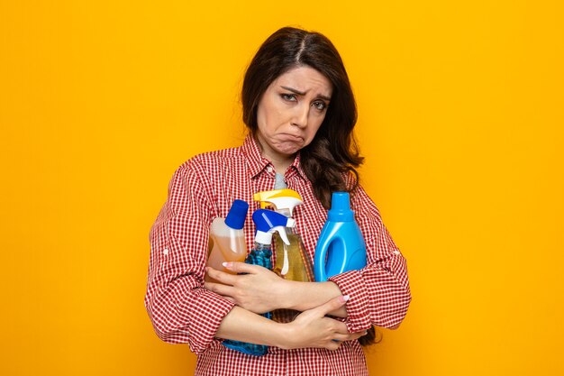 Sad pretty caucasian cleaner woman holding cleaning sprays and liquids 