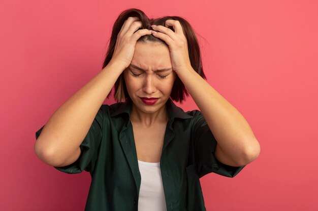 Sad pretty woman puts hands on head and pretends to cry isolated on pink wall