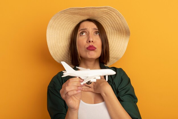 Sad pretty woman with beach hat holds model plane and looks up isolated on orange wall