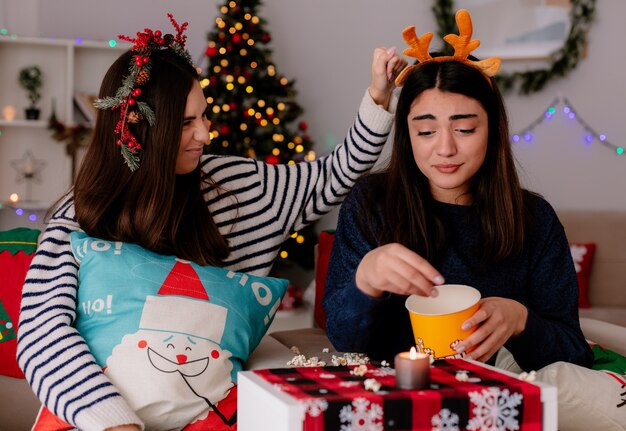 Sad pretty young girl with reindeer headband eats popcorn sitting on armchair with her friend christmas time at home