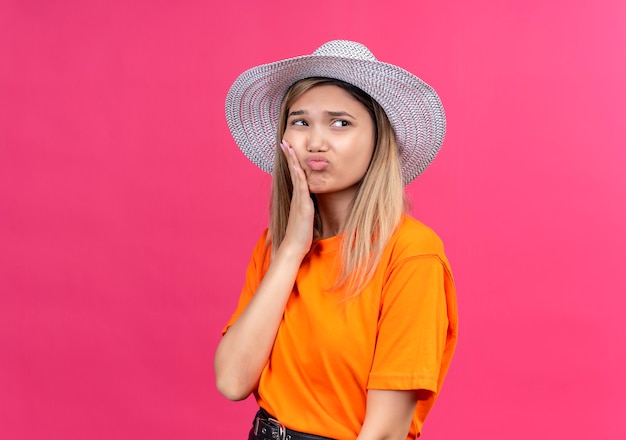 Una donna abbastanza giovane e triste in una maglietta arancione che indossa il cappello che tiene la mano sui suoi denti mentre ha dolore su una parete rosa