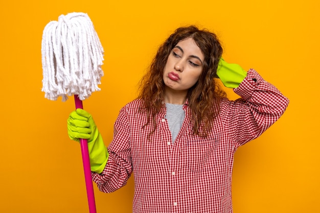 Sad putting hand on head young cleaning woman wearing gloves holding and looking at mop 