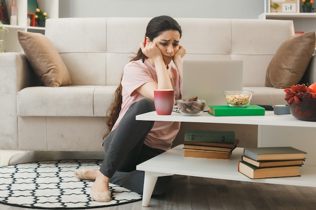 無料写真 悲しい頬に手を置く若い女の子は、リビングルームのコーヒーテーブルの後ろの床に座っているラップトップを使用しました