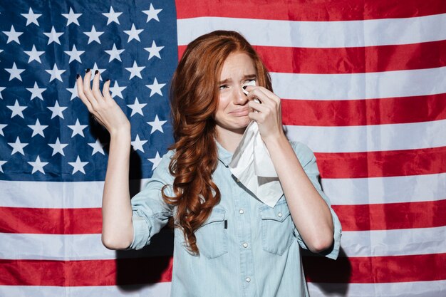 Sad redhead young lady cry over USA flag. Looking aside.