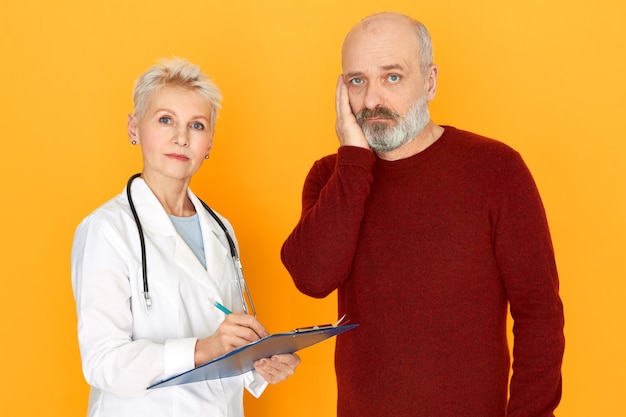 Sad retired female practitioner in white medical coat holding clipboard telling her elderly patient about diagnosis and treatment