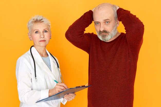 Sad retired female practitioner in white medical coat holding clipboard telling her elderly patient about diagnosis and treatment