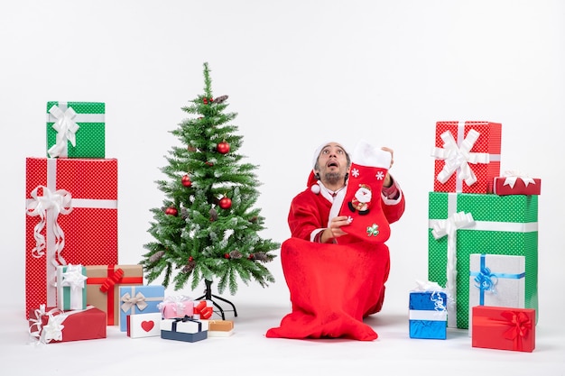 Sad santa claus looking above sitting on the ground and holding christmas sock near gifts and decorated new year tree on white background