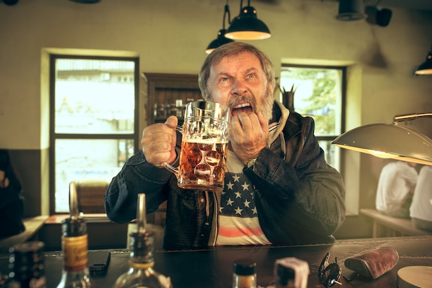 Печальный старший бородатый мужчина пьет пиво в пабе
