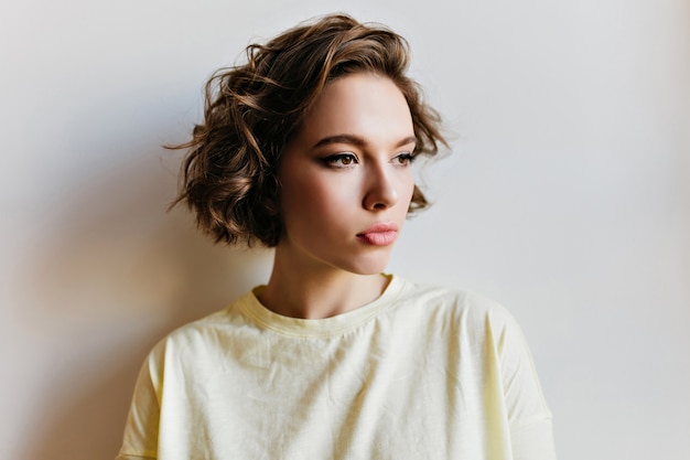 Sad short-haired girl with dark eyes looking away, standing on light wall. Close-up portrait of beautiful young woman with serious face expression thinking about something.