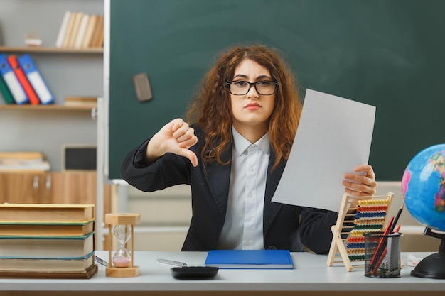 Triste che mostra i pollici verso il basso giovane insegnante femminile che indossa occhiali in possesso di carta seduto alla scrivania con strumenti scolastici in classe