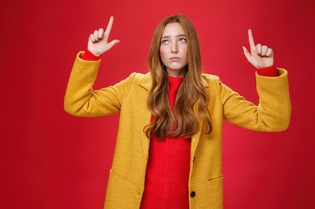 Triste e triste donna rossa carina in cappotto giallo alzando le mani scontento di rimpianto guardando l'angolo in alto a sinistra apatico come deluso dalla pioggia o dal maltempo sul muro rosso.