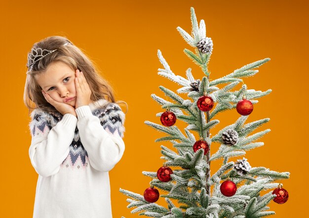 Sad tilting head little girl standing nearby christmas tree wearing tiara with garland on neck putting hands on cheeks isolated on orange background