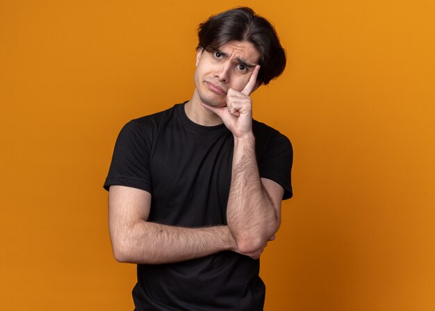 Sad tilting head young handsome guy wearing black t-shirt putting hand on cheek isolated on orange wall