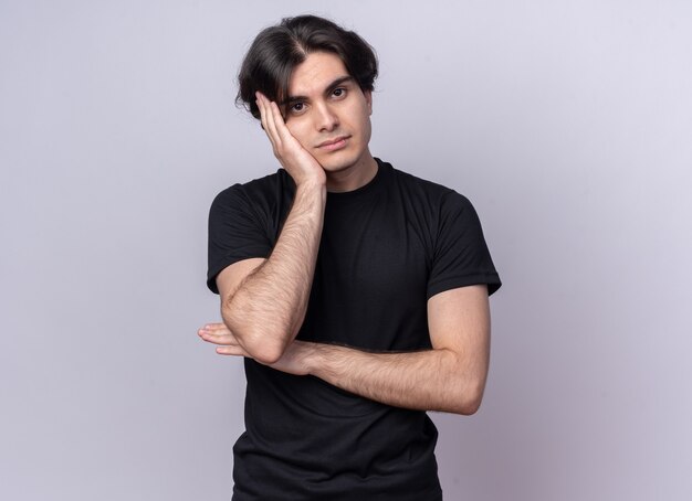 Sad tilting head young handsome guy wearing black t-shirt putting hand on cheek isolated on white wall