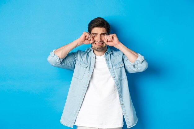 Sad timid man crying, wiping tears off face and sobbing, looking offended and upset, standing over blue background.