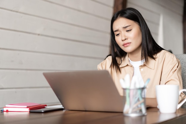 Sad and tired asian girl looking disappointed at laptop screen bored of studying watching something ...