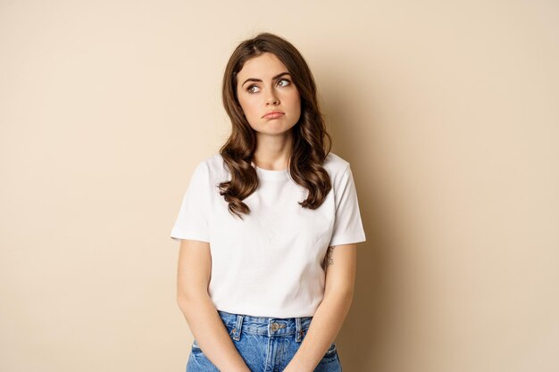 Sad and tired brunette woman looking drained and unamused standing with pokerface against beige back...