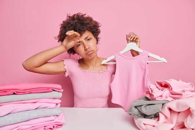 Sad tired young housewife sortes clothes for laundry holds pink shirt on hanger wipes forehead from tiredness sits at table with two stacks of clothing chooses something unnecessary for donation